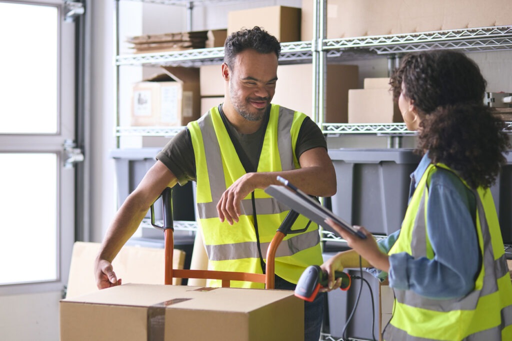 Woman Using Digital Tablet And Male Colleague With Down Syndrome Working In Warehouse